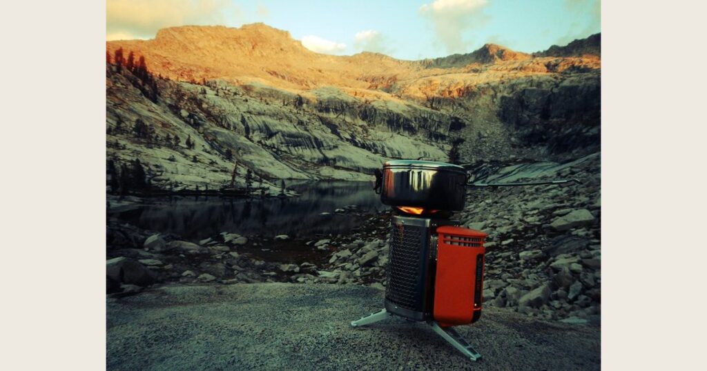 A black camp stove and red gear sit on rocks beside a mountain lake beneath sunlit ridges.